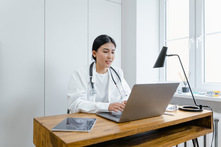 A female doctor in a white coat uses a laptop for an online consultation from her office