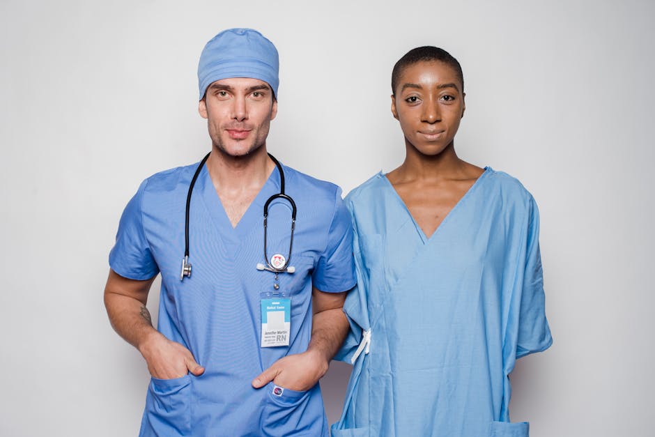 Positive male doctor in blue uniform with hands in pockets smiling and looking at camera while standing near nurse holding hands behind back in hospital